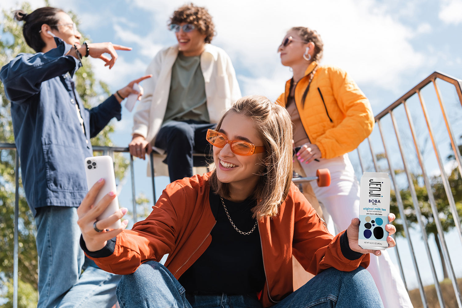 Woman taking a selfie with a can of Jenji Original Milk Tea she is holding while her skateboarding friends converse in the background