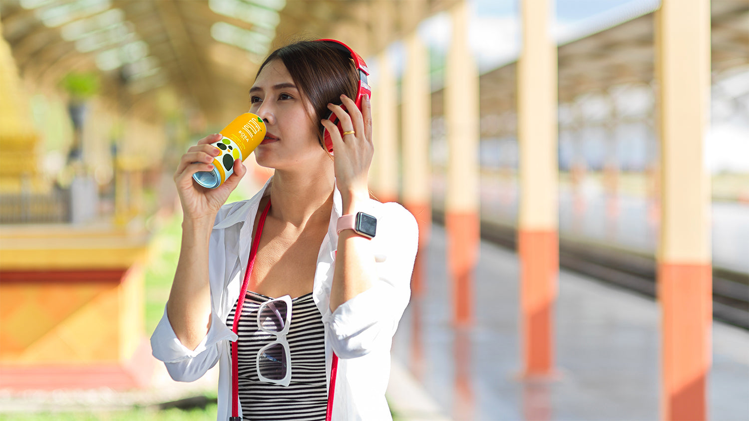 Young Asian woman listening to headphones at a train station on a leisure day drinking Jenji Pure Mango Boba Tea